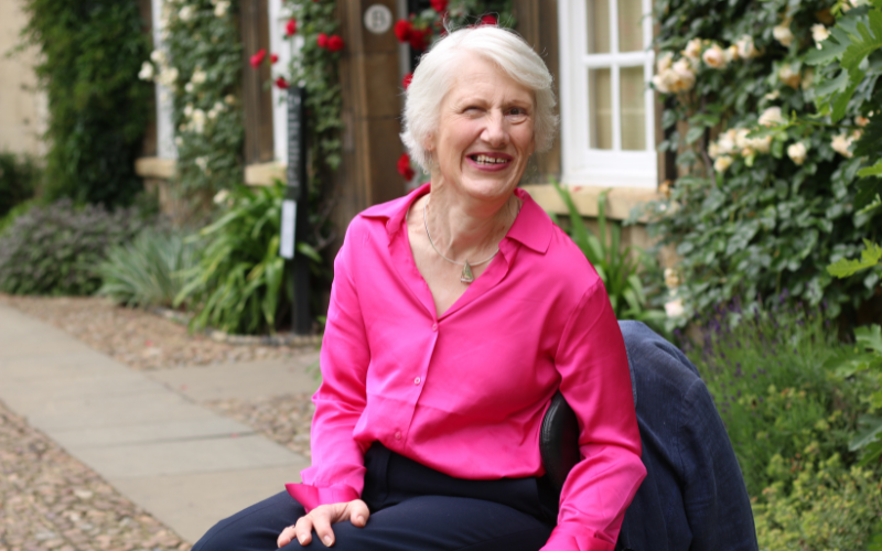 Sarah Stones wears a bright pink top, smiling widely in the flowery courtyard of a Cambridge College