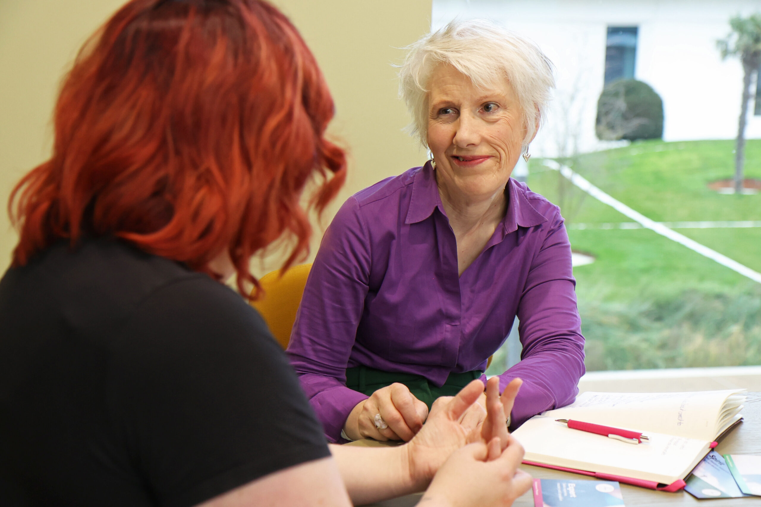 Two women are seated at a table engaged in conversation. One has red hair and is wearing a black shirt, while the other has short white hair and is dressed in a purple shirt. A notebook and pen are visible on the table, and the background features greenery outside a window.